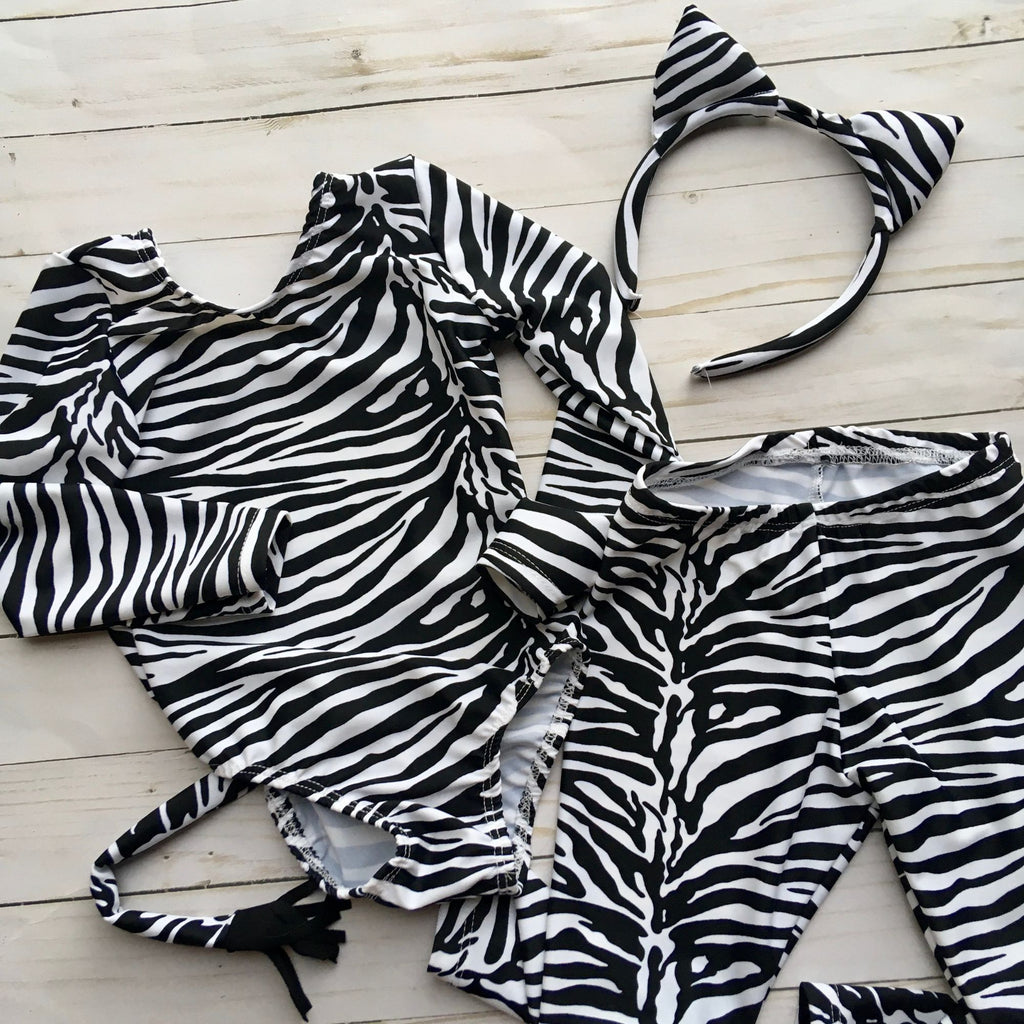 Zebra print, black and white stripe long sleeve leotard with attached tail with black fringe detail at end.  Athletic stretch fabric for second skin fit. Matching zebra print leggings and ear headband shown
